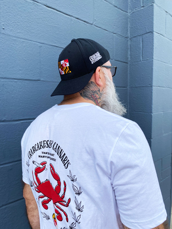 Man wearing black crabcakes and cannabis snapback and white crab emblem tee shirt, standing on a grey wall.
